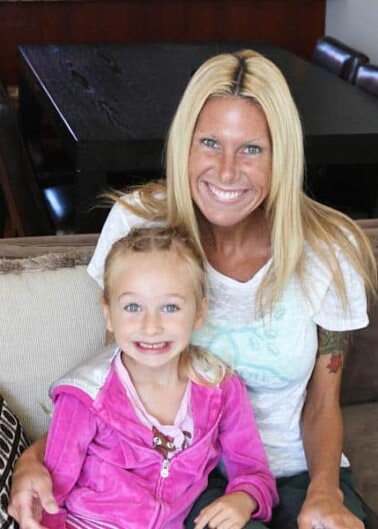 A smiling woman with blonde hair and a child in a pink jacket sitting on a sofa indoors.