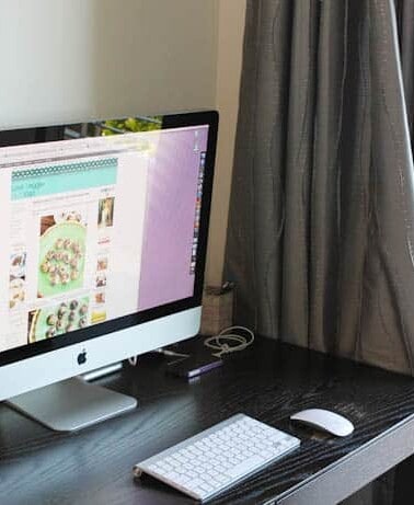 A desktop computer with an open application on the screen, a keyboard, and a mouse on a black desk against a curtained window.