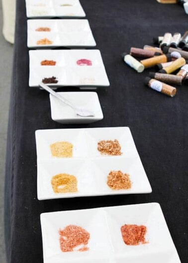 Assorted spices displayed on small white plates arranged on a black tablecloth, with spice bottles lying beside them.