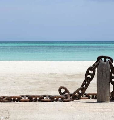 A large, rusty anchor chain stretches across a sandy beach with a tranquil blue sea in the background.