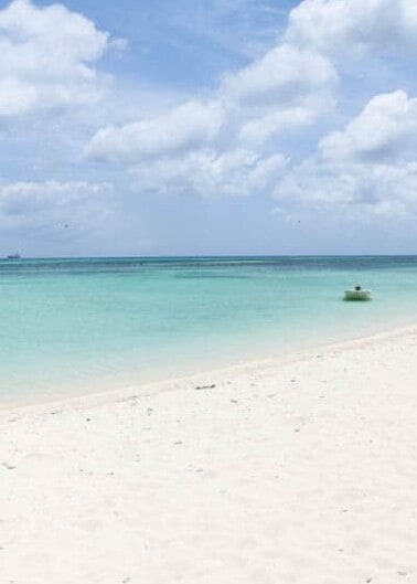 Tranquil beach scene with clear turquoise waters, white sand, and a lone person floating on the horizon.