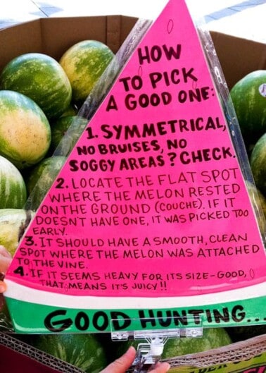 A person holding a pink sign with tips on how to pick a good watermelon, with watermelons in the background.