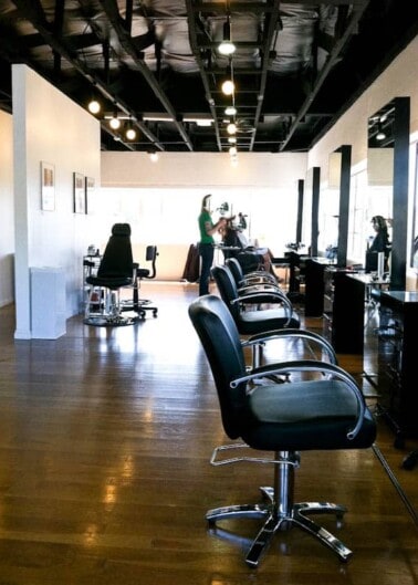 Empty chairs in a well-lit hair salon with a stylist working in the background.