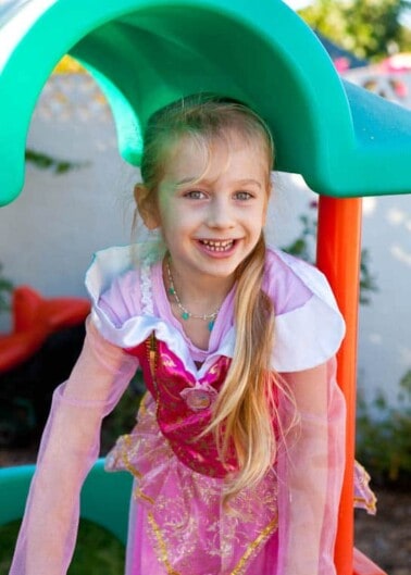 A smiling girl in a pink princess dress playing under a play structure.