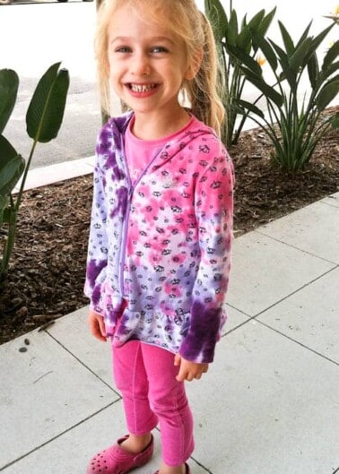 A young girl smiling with missing front teeth, wearing a purple patterned jacket, pink pants, and pink crocs, standing on a sidewalk with plants in the background.