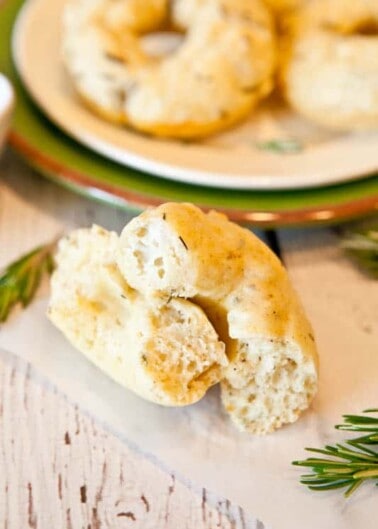 A close-up of a broken biscuit with more biscuits on a plate in the background, on a wooden surface with a sprig of rosemary.