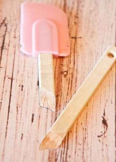 A pink ice cream bar melting on a wooden surface with the stick laid beside it.