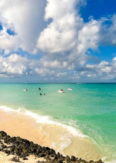 Tropical beach with clear turquoise waters, sandy shore, and scattered clouds in the sky.