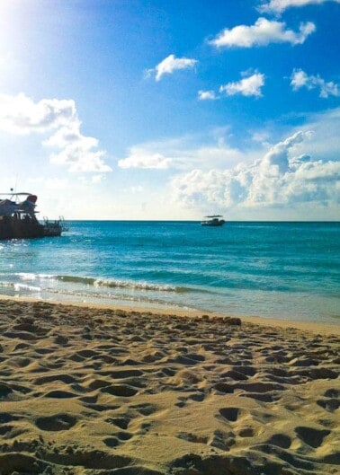 Sunny beach scene with clear blue water, boats, and footprints in the sand.