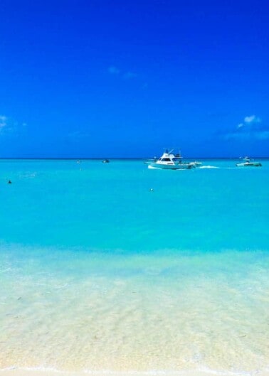 A tranquil beach scene with clear turquoise waters, a white sand shore, and boats floating near the horizon under a bright blue sky.