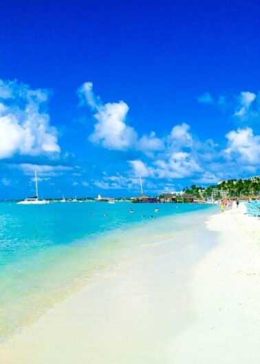 A tropical beach with clear blue skies, white sand, and a few boats on the horizon.