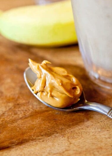 A spoonful of peanut butter on a wooden surface with a banana and a glass of smoothie in the background.