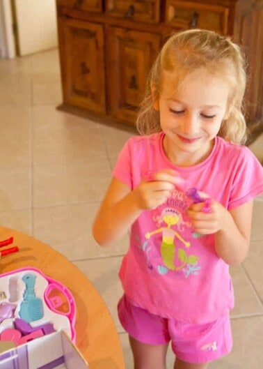 A young girl in a pink shirt smiling while playing with toys in a room.