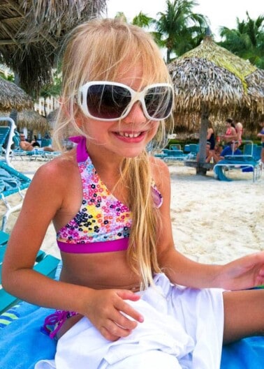 A young girl wearing oversized sunglasses and a colorful swimsuit sits on a beach chair, with palm-thatched umbrellas and beachgoers in the background.