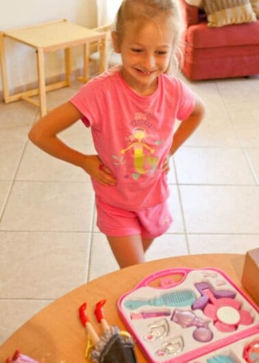 Young girl standing with hands on hips, smiling at a selection of toy beauty salon tools on a table.