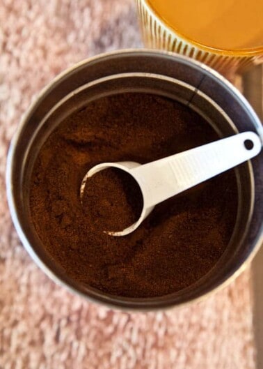 A white measuring spoon in a container of ground coffee with a glass of orange juice in the background.