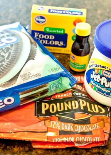 Ingredients for a baking recipe consisting of oreos, assorted food coloring, marshmallow creme, and a bar of dark chocolate on a kitchen counter.