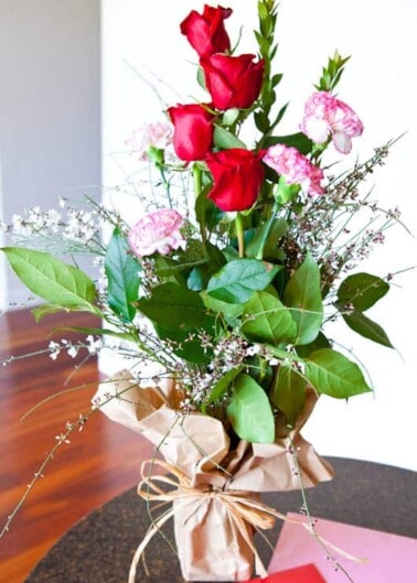 A bouquet of red roses and pink carnations wrapped in brown paper on a table.