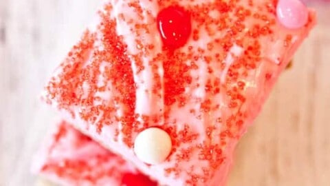 A piece of pink frosted cake with red sprinkles and candy toppings on a wooden surface.