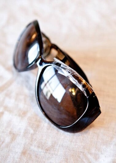 A pair of broken sunglasses with one lens taped up lying on a textured surface.