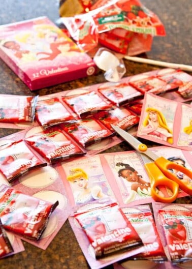 Assorted disney-themed valentine's day cards and candies spread out on a table with scissors.