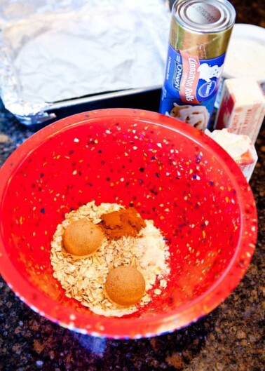 Ingredients for a recipe in a red mixing bowl, including oats and spices, with canned goods and butter nearby on the counter.