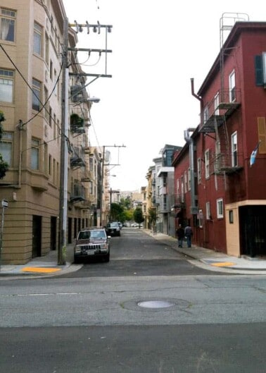 A steep urban street lined with multistory buildings and parked cars, with a pedestrian walking on the sidewalk.