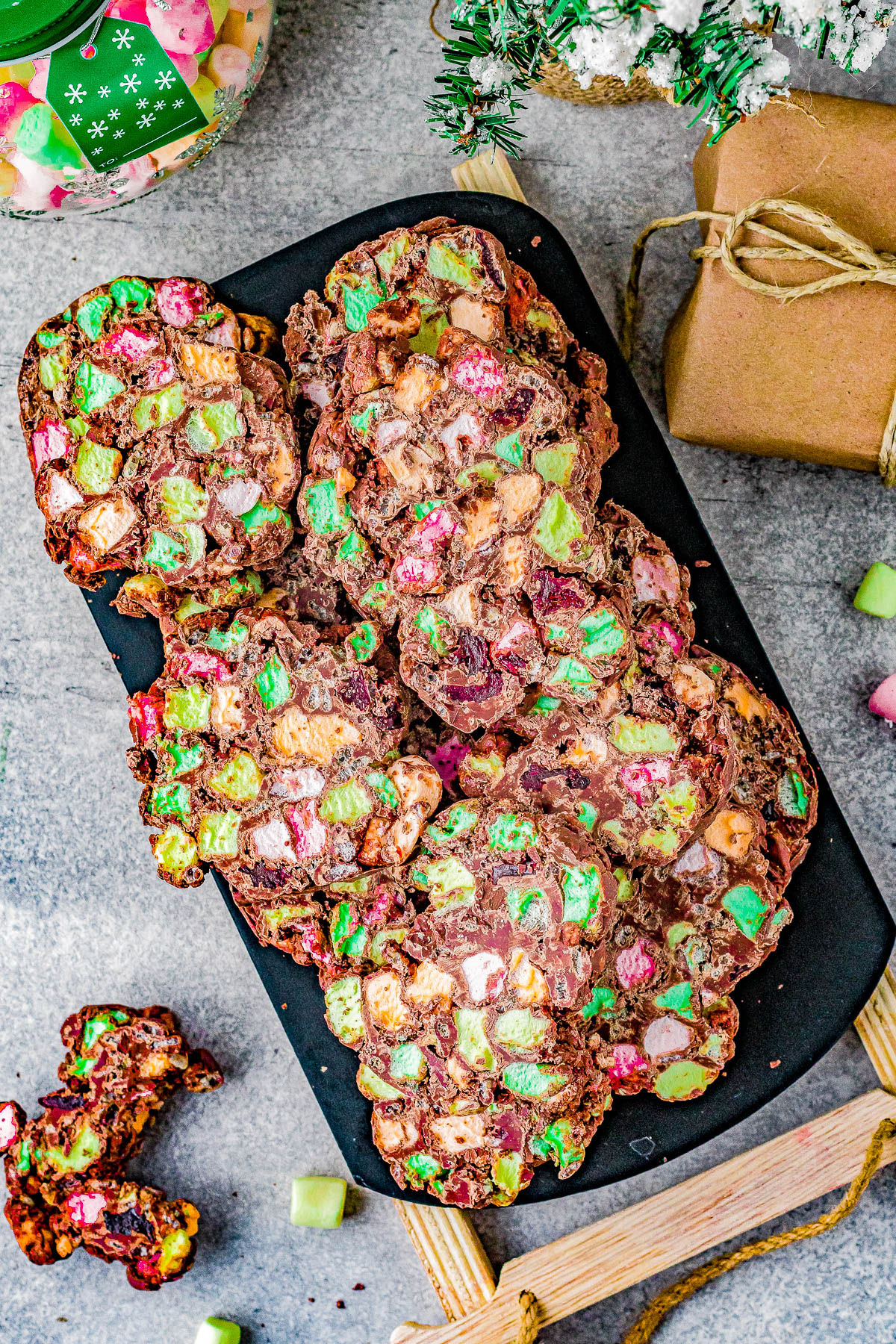 Colorful rocky road slices with marshmallows and chocolate are displayed on a dark serving board. A wrapped gift and a jar of treats are nearby on a gray countertop.