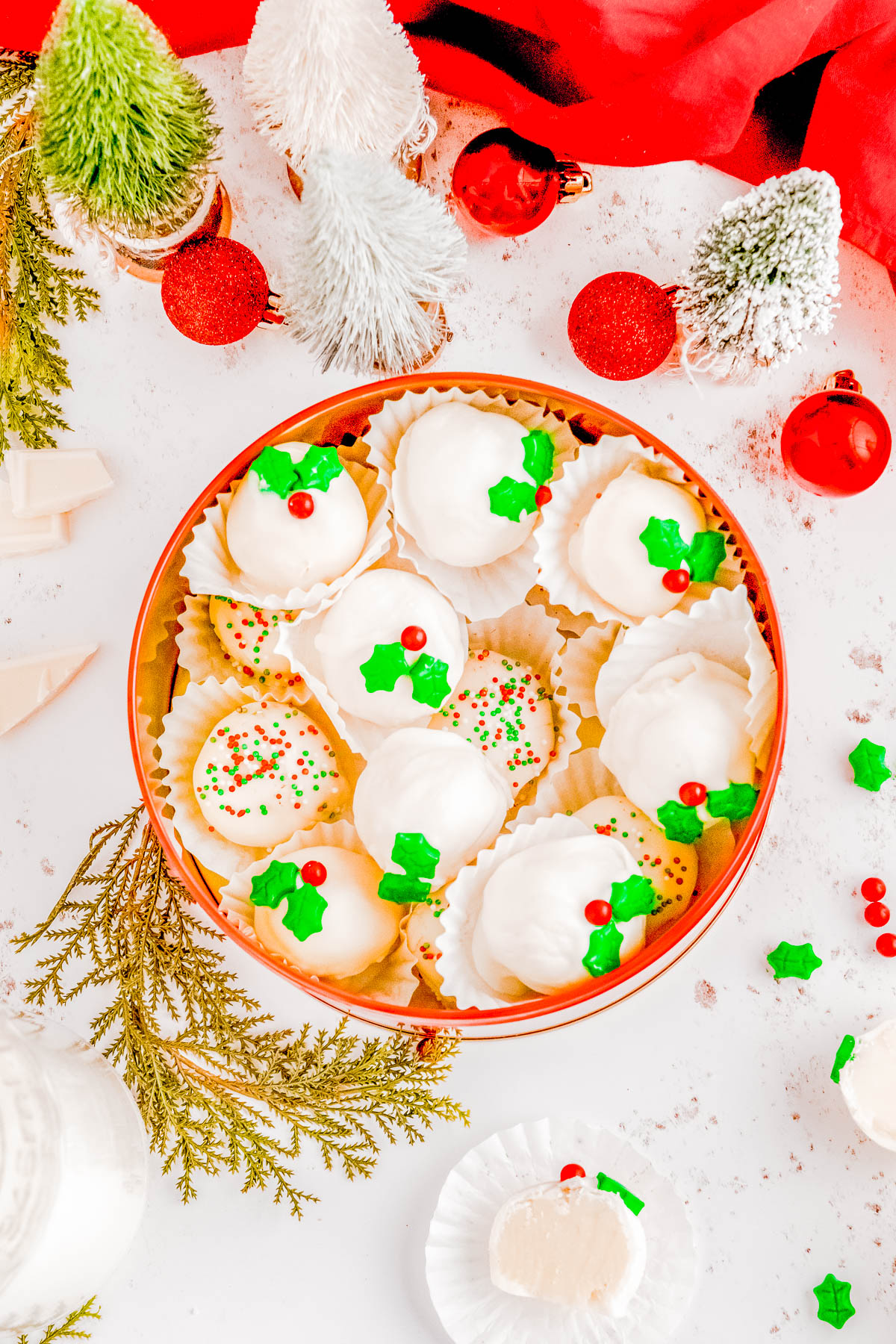 A tin of decorated white holiday cookies with green and red accents, surrounded by small Christmas ornaments and decorative trees.