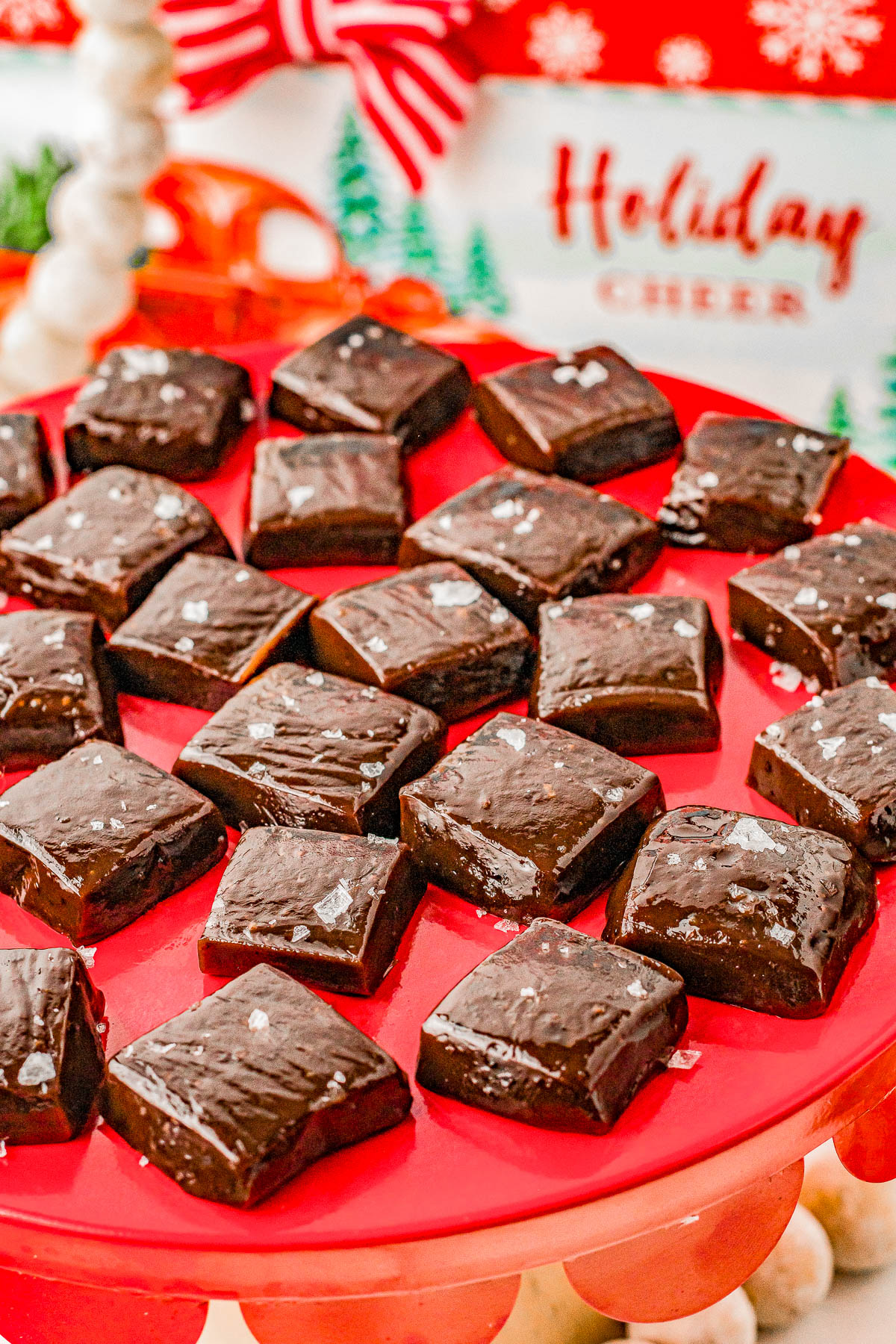 Squares of chocolate fudge with a sprinkle of sea salt are arranged on a red plate. Holiday-themed decorations are visible in the background.