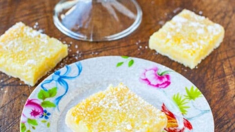 Lemon bars dusted with powdered sugar on a decorative plate with more in the background on a glass stand.