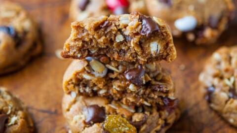 A variety of freshly baked cookies with chocolate chunks and nuts, one broken in half to reveal the chewy interior, on a wooden surface.