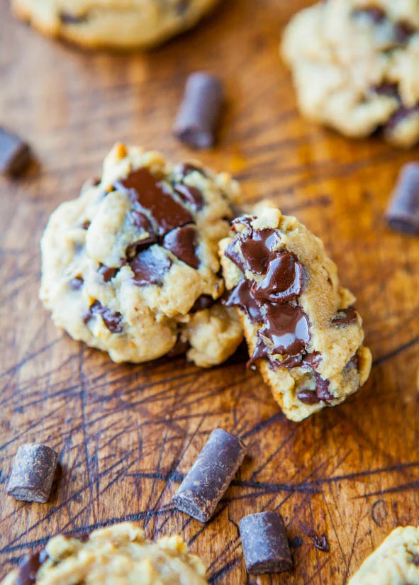 Close-up of chocolate chip cookies on a wooden surface, with melted chocolate visible. Chocolate pieces are scattered around.