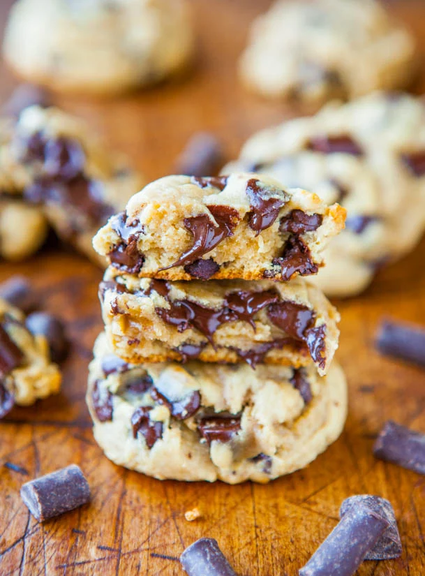 Close-up of chocolate chip cookies with melted chocolate, stacked on a wooden surface with chocolate chunks scattered around.