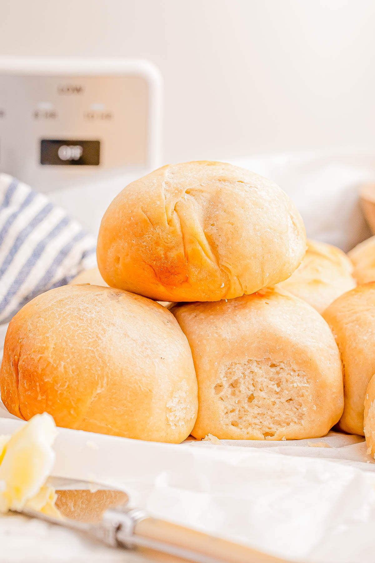 A stack of freshly baked dinner rolls on a white surface, with a butter knife and a slice of butter nearby.