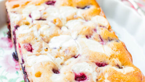 A freshly baked blueberry loaf cake with a sugar glaze, presented on a decorative plate with a pink and white checked cloth beneath it.