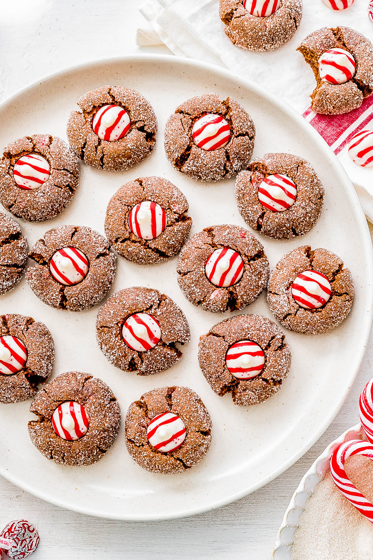 A plate of freshly baked chocolate cookies topped with red and white striped peppermint candies. Additional cookies and a bowl of sugar are nearby.