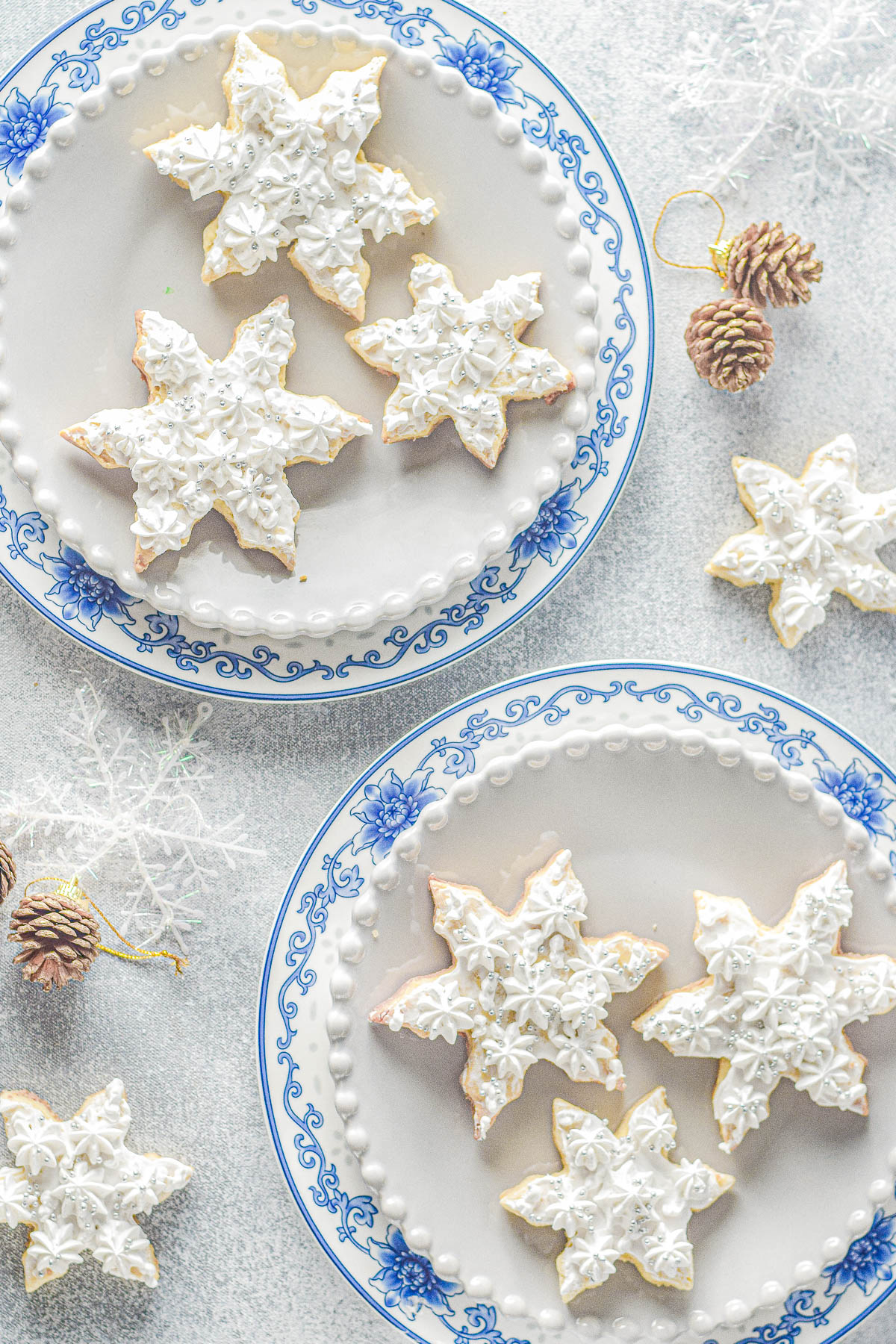 Two plates of snowflake-shaped cookies with white frosting, set on a light background with decorative pine cones and snowflakes.