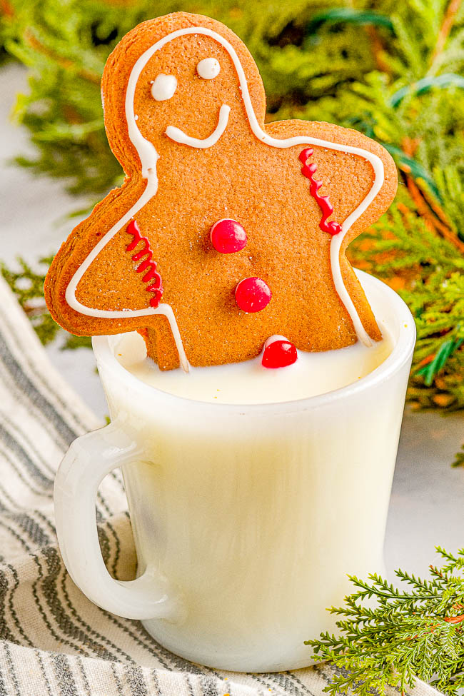 A gingerbread cookie with icing is placed on the rim of a white mug filled with milk, set against a background of greenery and a fabric cloth.