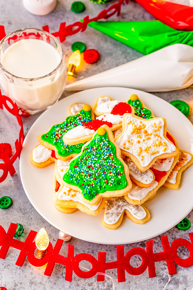 A plate of decorated Christmas cookies in the shape of trees and stars, with a glass of milk beside them. Festive decorations are scattered around.