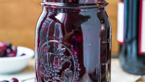 A glass jar filled with dark purple fruit jam, topped with whole berries and red wine cranberry sauce, on a wooden table.