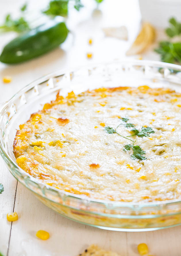 A round, clear glass dish containing a baked casserole topped with melted cheese and garnished with cilantro sits on a white surface, surrounded by scattered corn kernels and a green pepper in the background.
