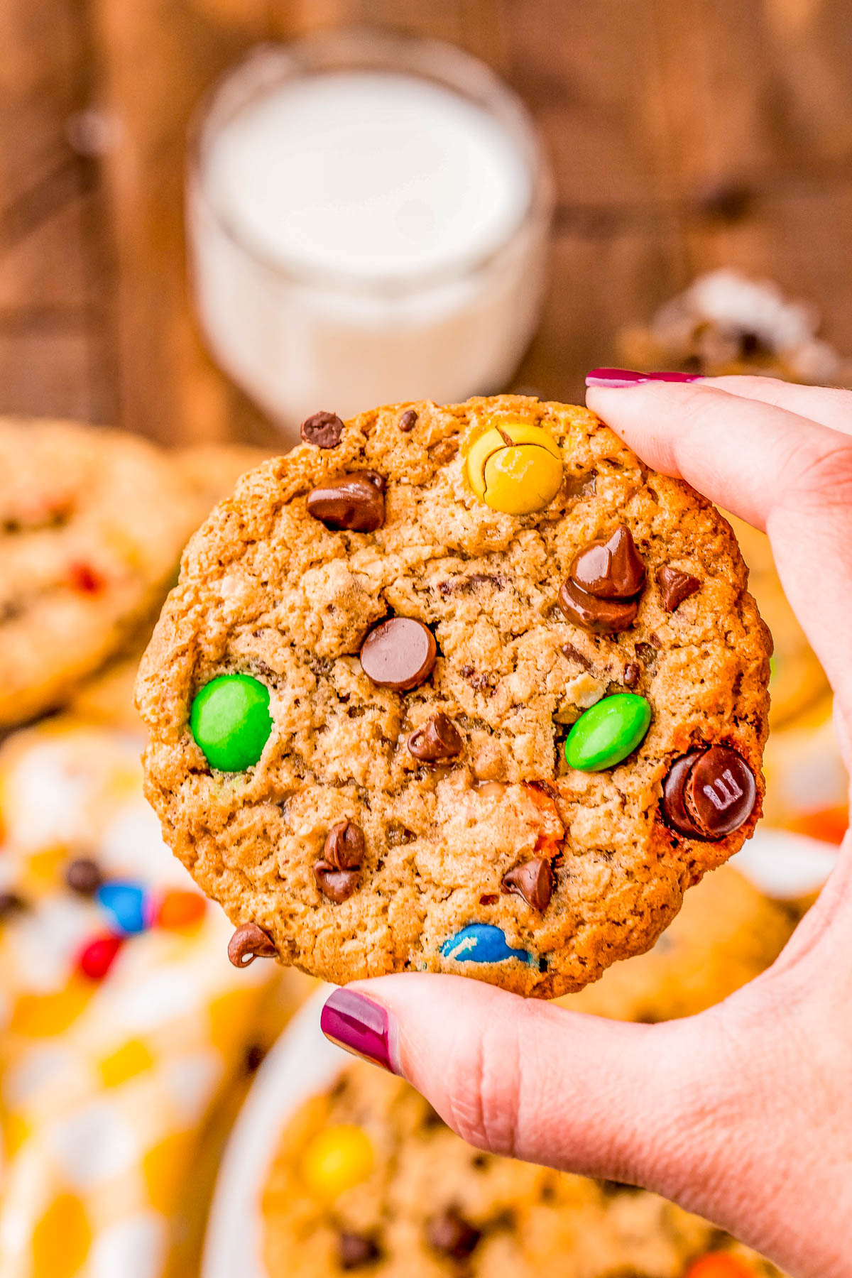 Hand holding a cookie with chocolate chips and colorful candies, with a glass of milk in the background.