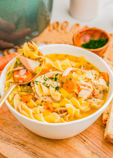 A bowl of chicken noodle soup with vegetables and herbs on a wooden board, accompanied by slices of bread and a small bowl of chopped parsley.