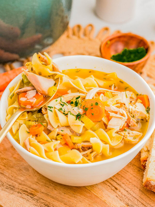 A bowl of chicken noodle soup with vegetables and herbs on a wooden board, accompanied by slices of bread and a small bowl of chopped parsley.