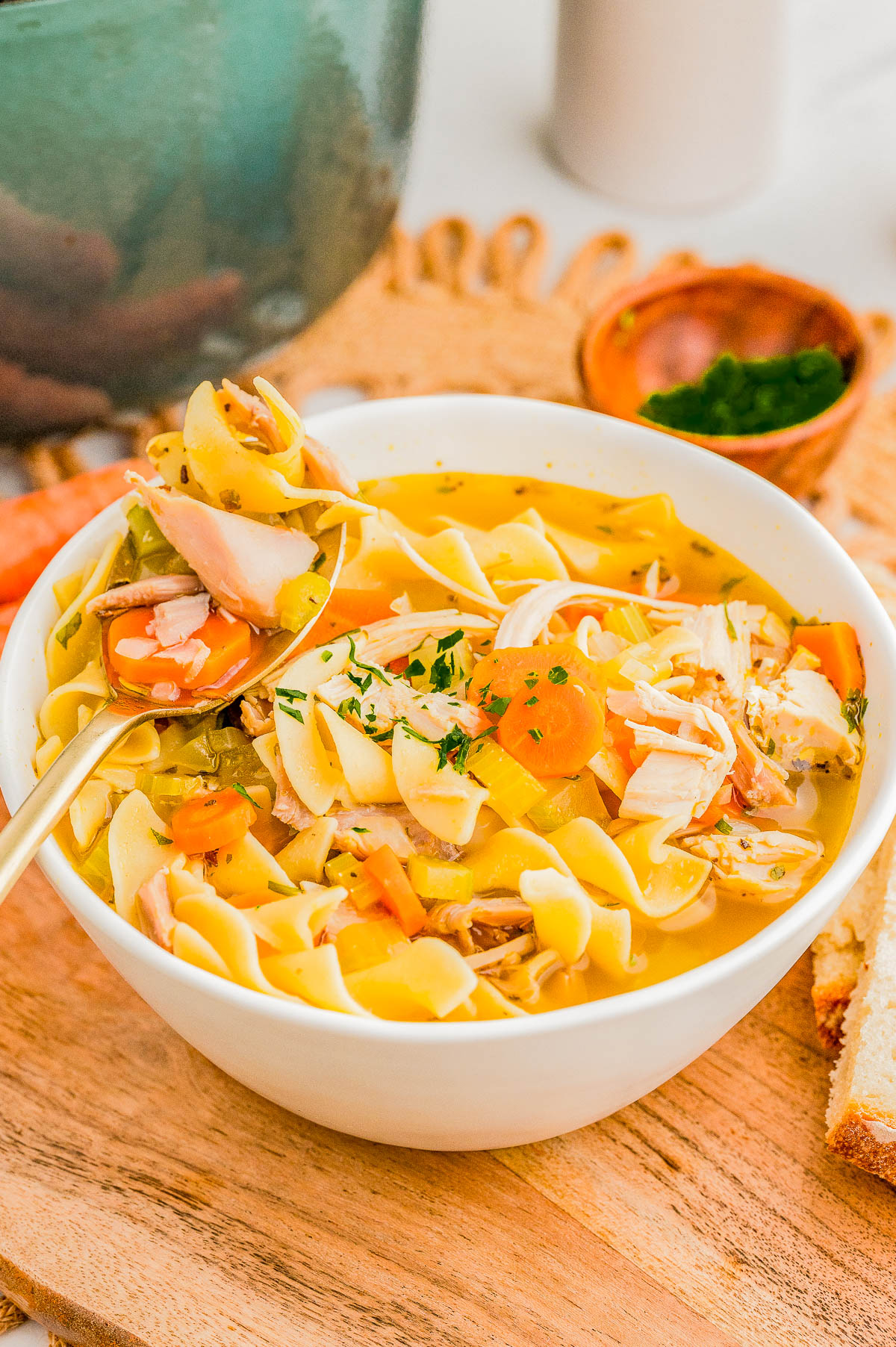 A bowl of chicken noodle soup with vegetables and herbs on a wooden board, accompanied by slices of bread and a small bowl of chopped parsley.