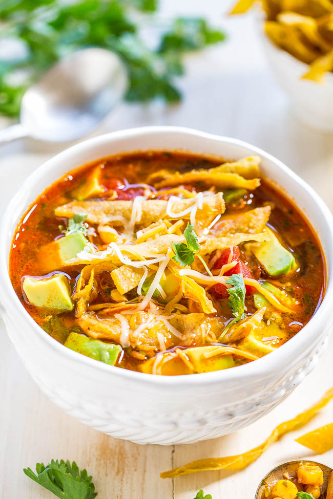 A bowl of chicken tortilla soup garnished with avocado slices and cheese, served on a wooden table.