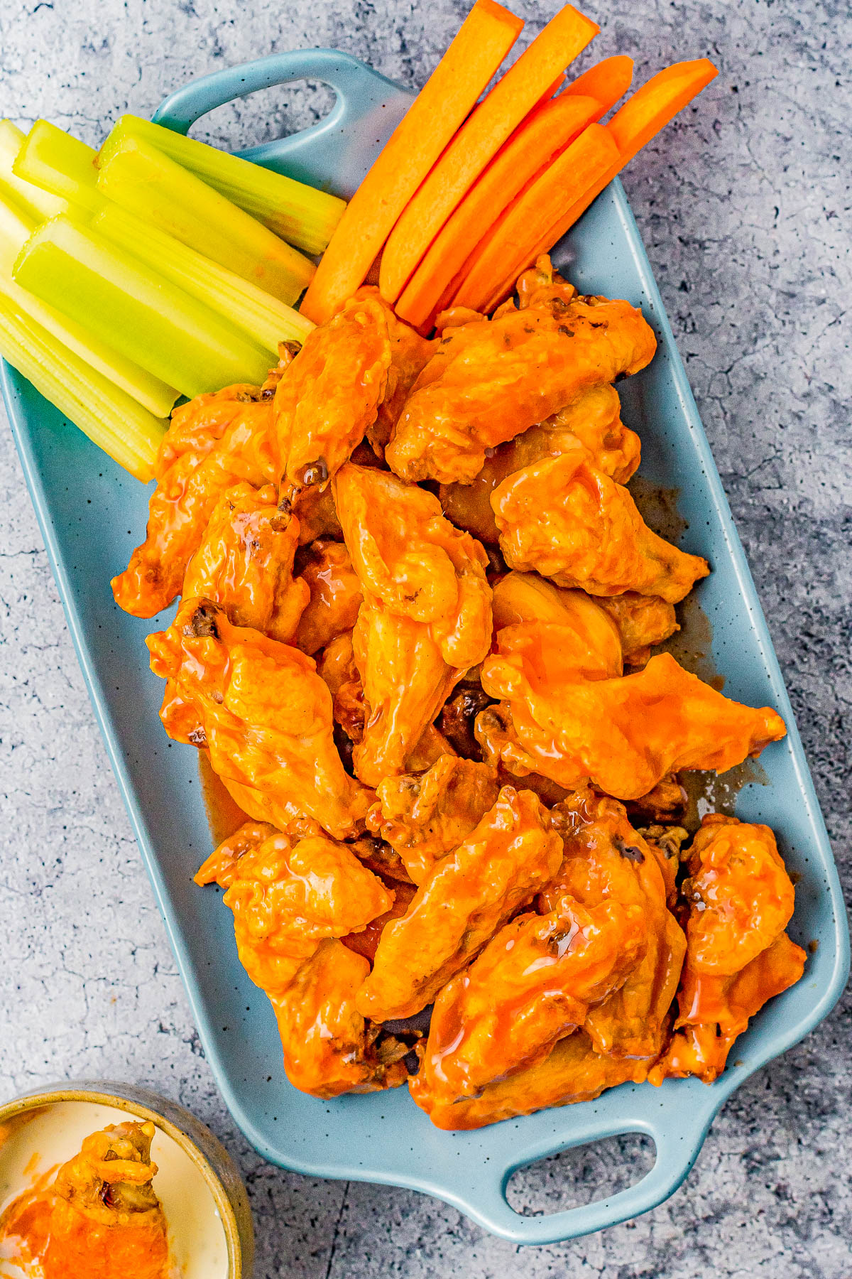 A platter of buffalo chicken wings with carrot and celery sticks on the side, accompanied by a dipping sauce in a dish on a gray surface.