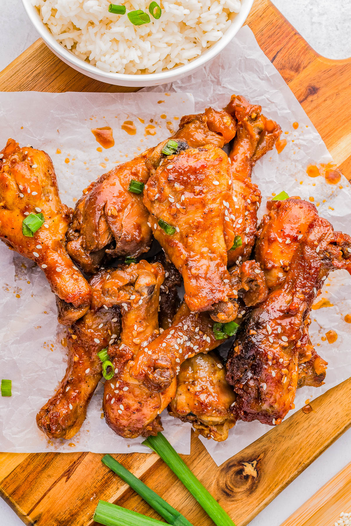 A pile of glazed chicken drumsticks garnished with sesame seeds and green onions on parchment paper, with a bowl of rice in the background.