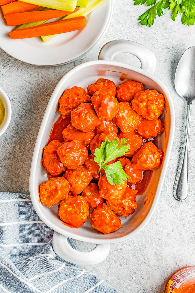 Buffalo cauliflower bites in a white dish, garnished with parsley, next to a plate of carrot and celery sticks. A spoon rests on the gray surface.